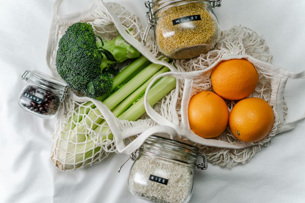 Oranges, celery, and broccoli on a table with millet, beans, and rice in separate glass jars with lids that are all a part of a special diet and exercise routine