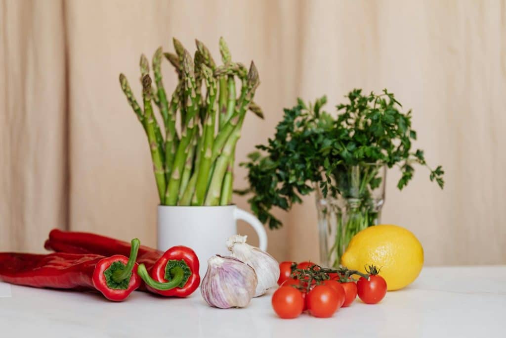 Composition of red cherry tomatoes and peppers with garlic and yellow lemon near a white cup with asparagus and green parsley in a glass on the table